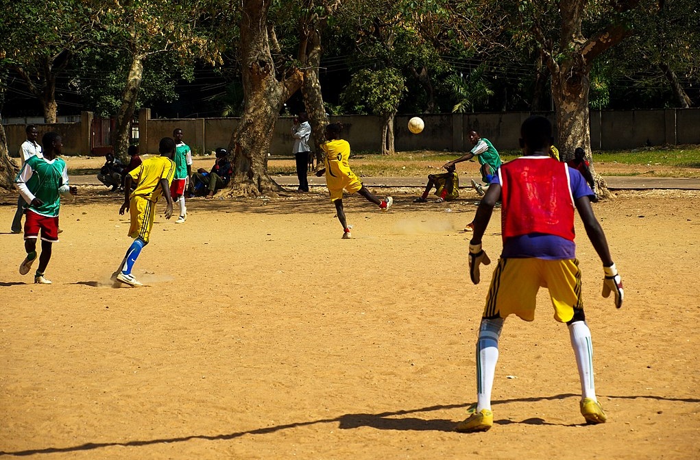 Men in red and yellow strips playing football on sand.