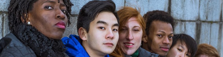 A black woman, asian man, white redhead woman, black man, and asian woman sitting against a grey brick wall