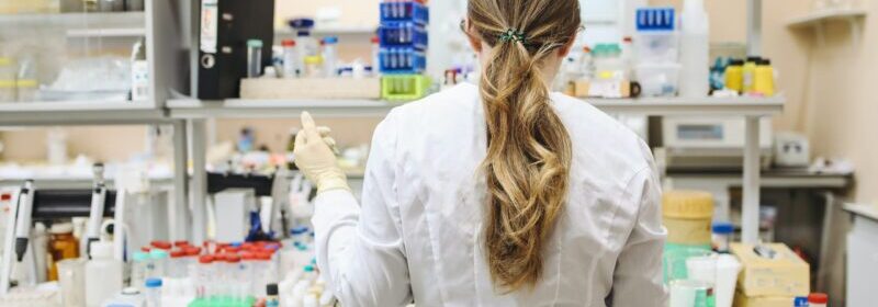 Woman in lab coat stands in front of a multi tiered desk filled with test tubes, beakers and lab equipment